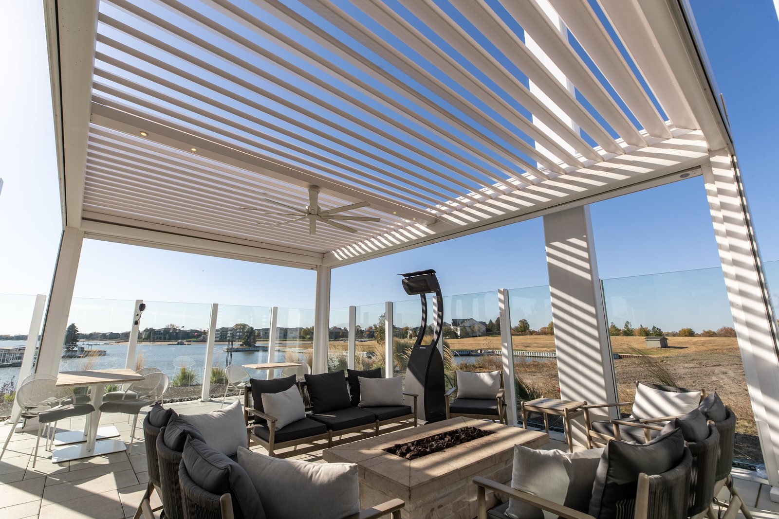 White Azenco R-BLADE motorized louvered pergola with recessed lighting, ceiling fan, patio heater and firepit lounge seating overlooking a lake